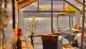 Una habitación con un ventanal grande, una cama, una silla, una mesa y una mesita de noche con un jarrón de flores.