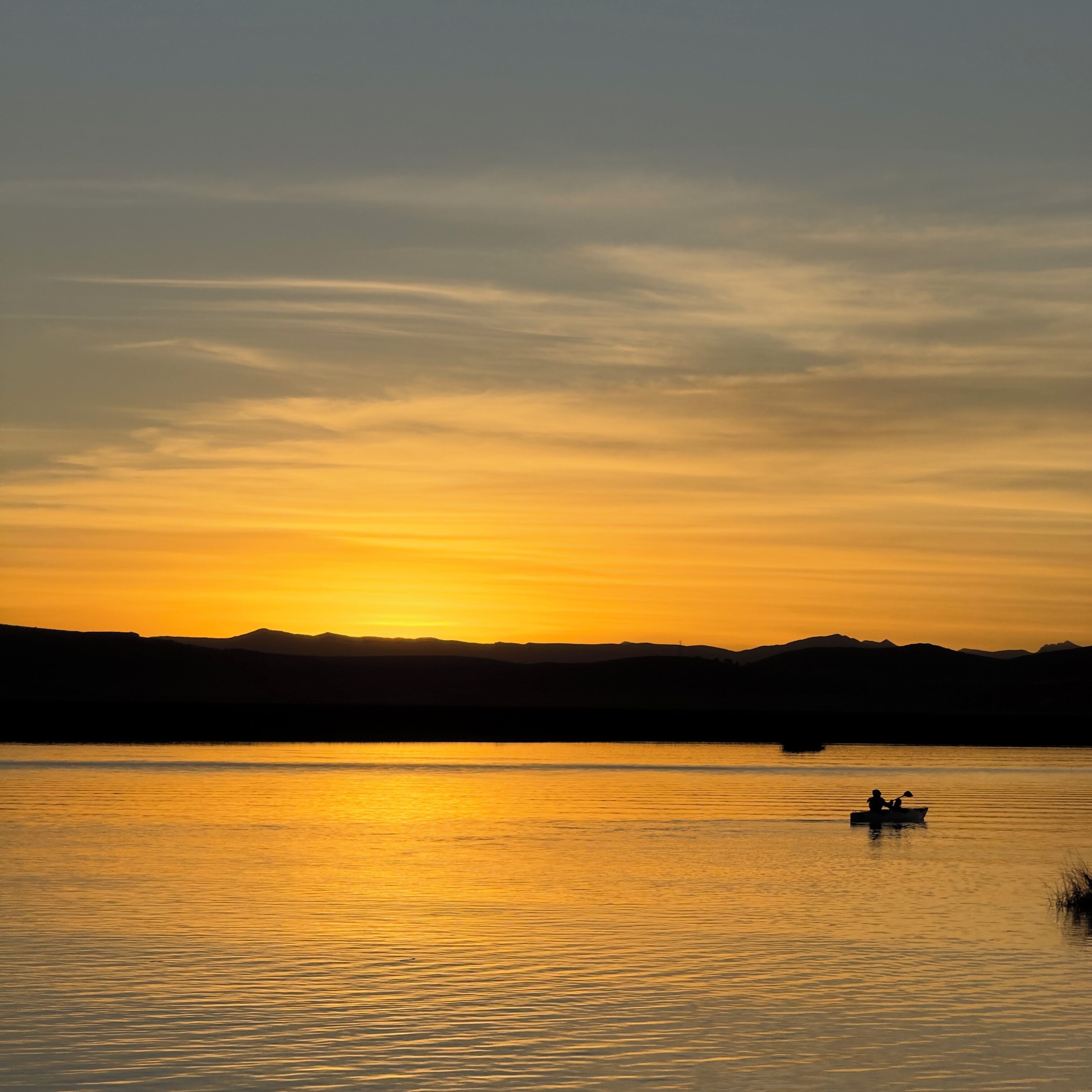 A calm lake at sunset with a silhouette of a boat and distant mountains.