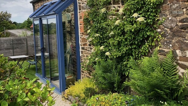 Un bâtiment en pierre doté d’une véranda en verre, entouré d’un chemin de gravier et de diverses plantes.