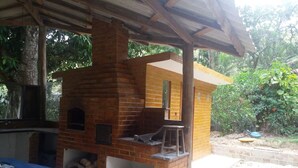 A brick-built outdoor kitchen with a chimney, a table with a blue tablecloth, and a garden area with plants and a tree. - SITE REFUGIO DO RIO - BANANAL - SP (BANANAL)