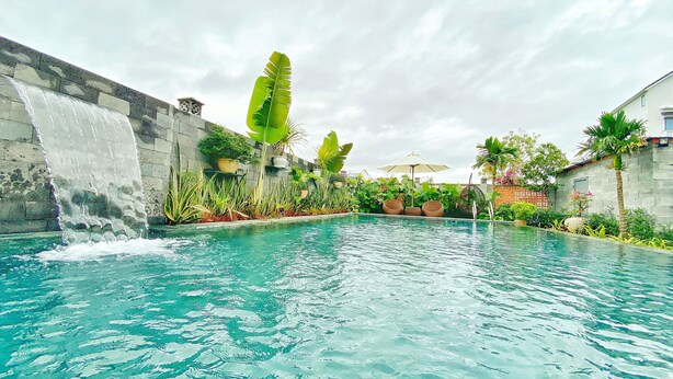 A swimming pool with a waterfall feature, surrounded by lush greenery and potted plants. - Hoi An Cabbage Garden (Da Nang)
