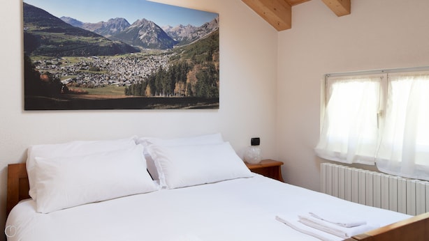 Un dormitorio con una cama grande, ropa de cama blanca, una mesita de noche con lámpara, una ventana con cortinas translúcidas y un cuadro de un paisaje montañoso en la pared.