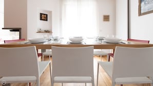 Un coin repas avec une table en bois dressée pour quatre personnes, des chaises blanches et un mur blanc orné d’un tableau et d’une étagère.