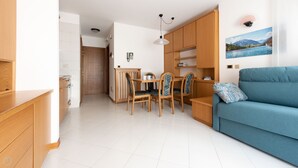 A modern living room with a blue sofa, wooden dining table with chairs, and a kitchen area with wooden cabinets. - San Giovanni 13/A - Foliage (Molina)