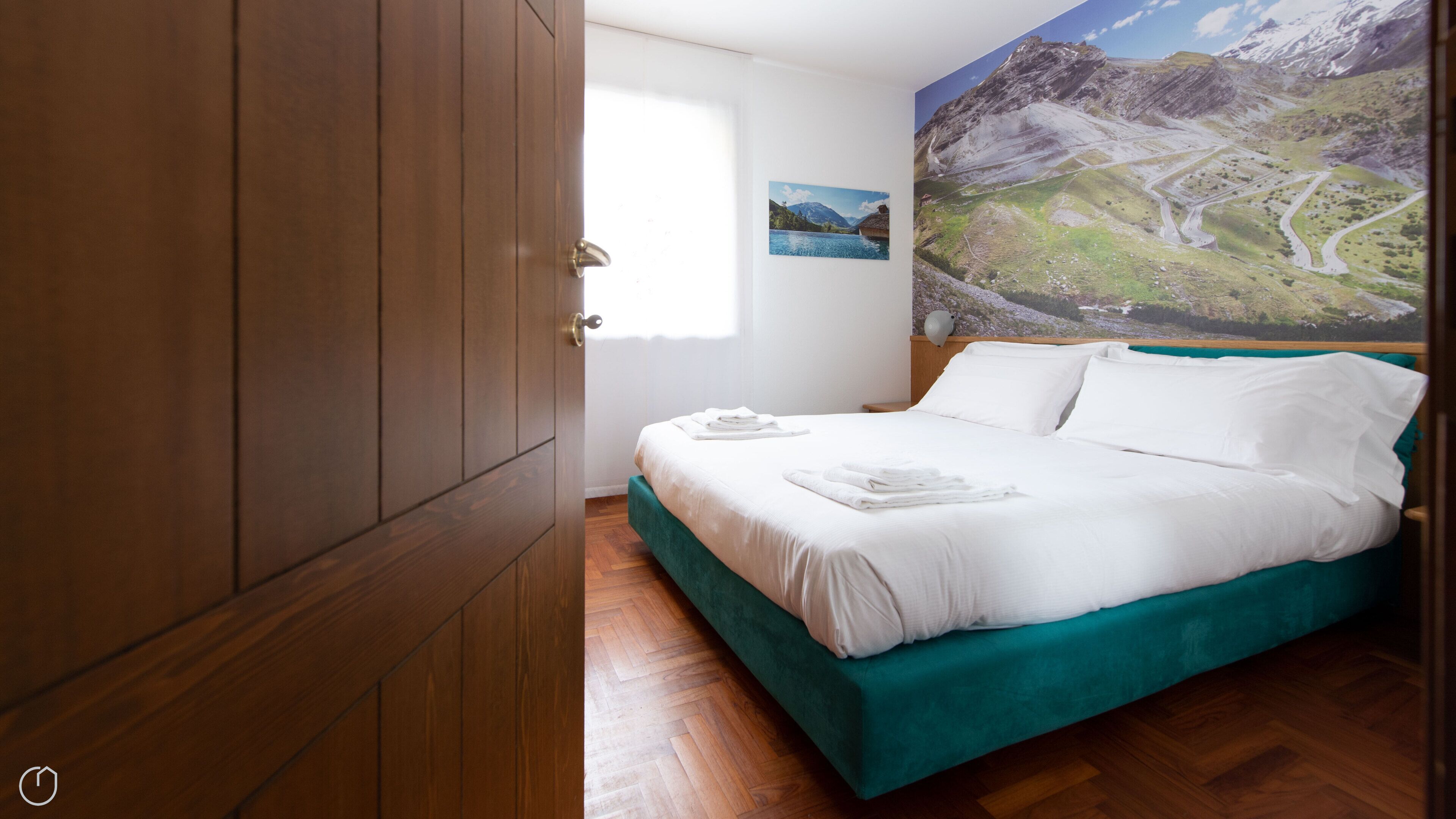 A bedroom with a wooden wardrobe, a bed with white linens, a green headboard, and a large landscape painting on the wall.