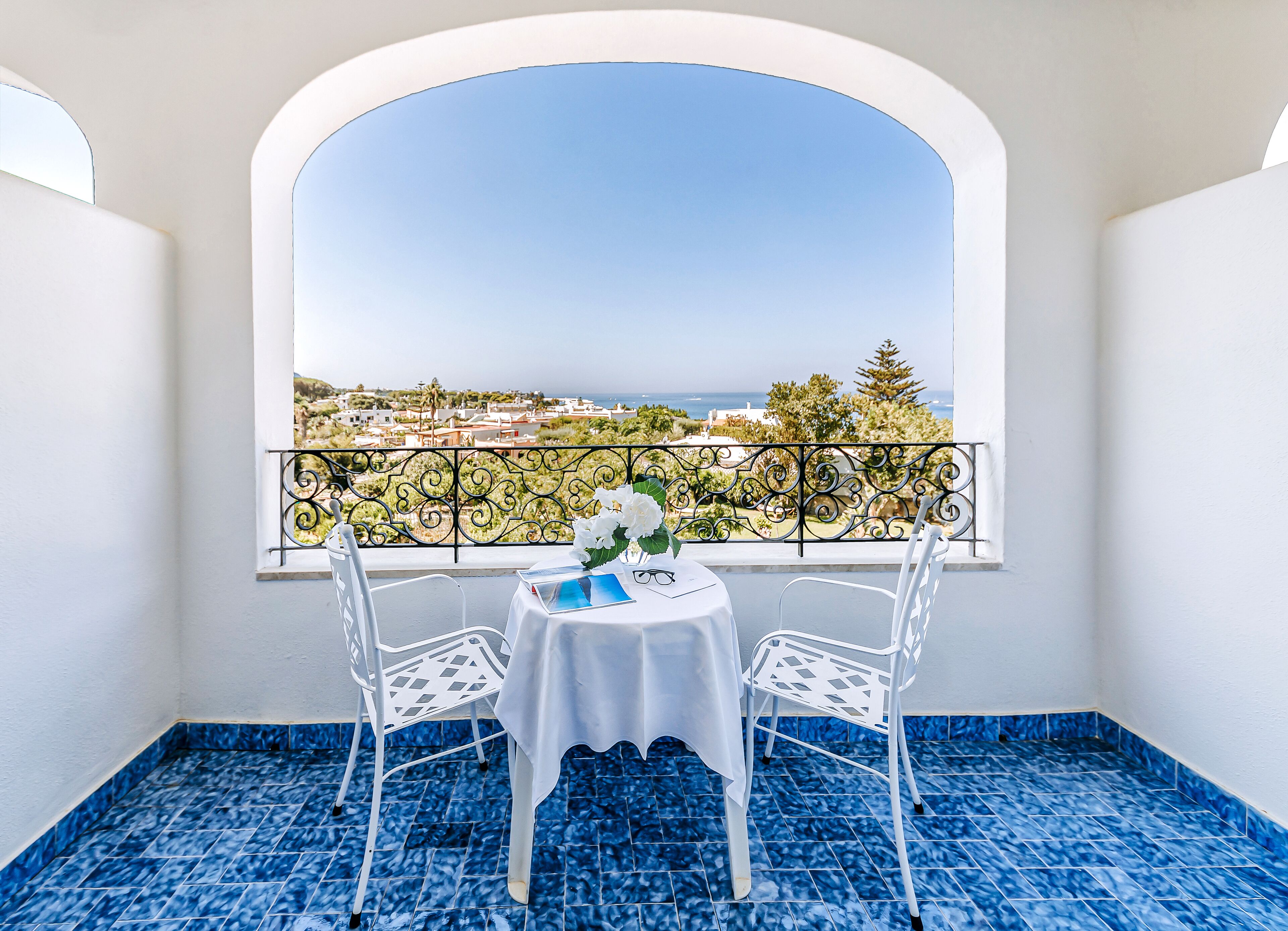 Un balcone con tavolo e sedie, vista su una cittadina costiera e pavimento con piastrelle a motivi blu.