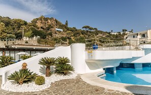 Zona de piscina con una estructura blanca, palmeras y vista a una ladera con edificios.