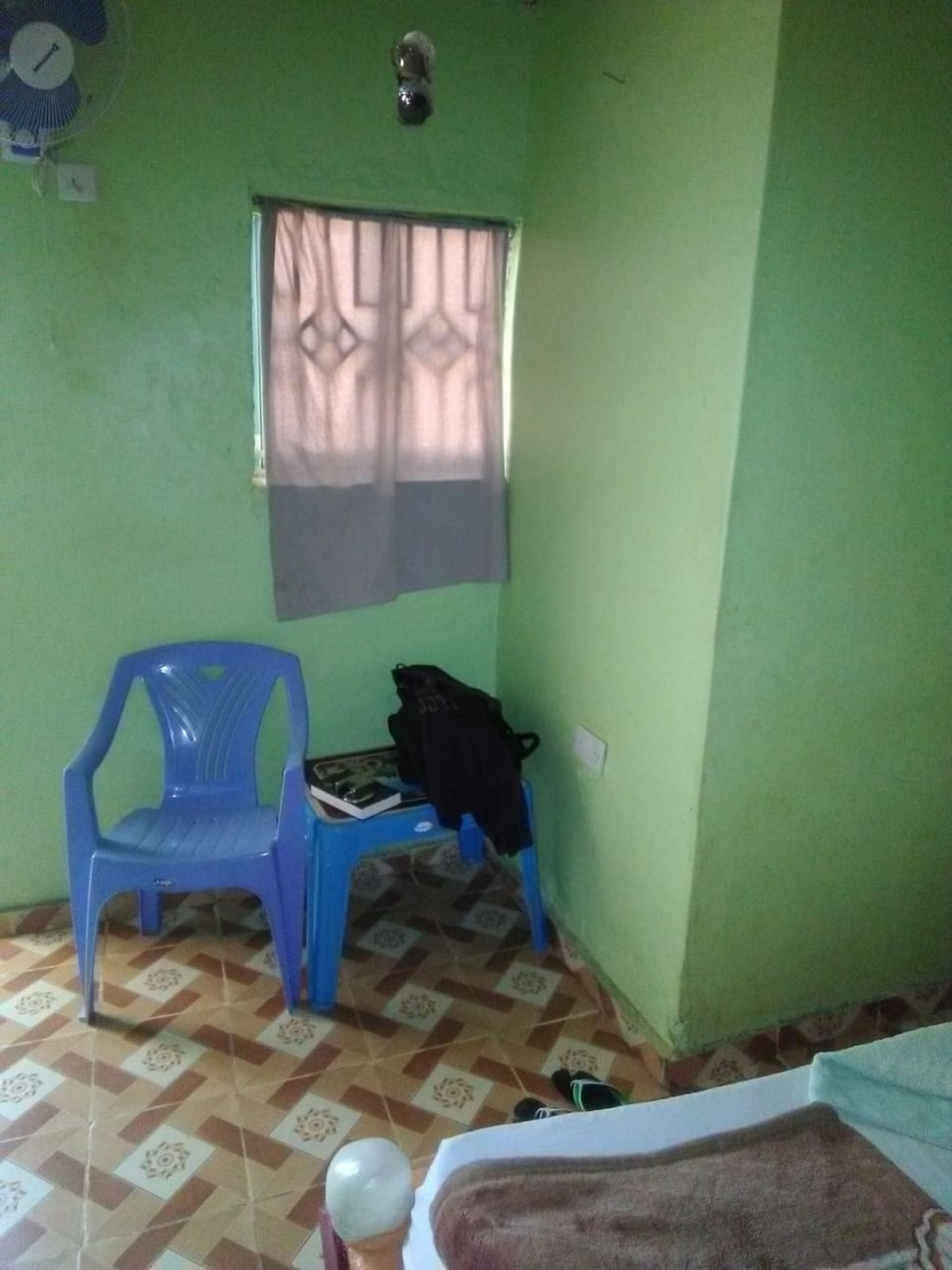 Una habitación con paredes verdes, una ventana con cortina, una silla de plástico azul, una mesa pequeña con un bolso negro y un suelo estampado.