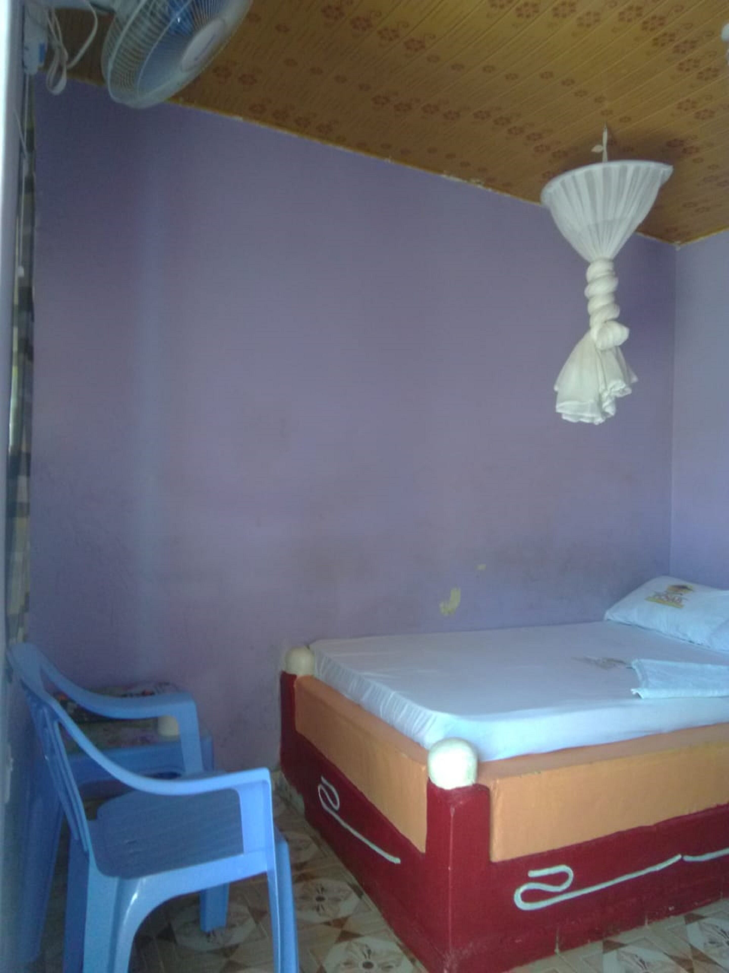 Una habitación pequeña con cama individual, cabecera de madera, una silla de plástico azul y ventilador de techo.