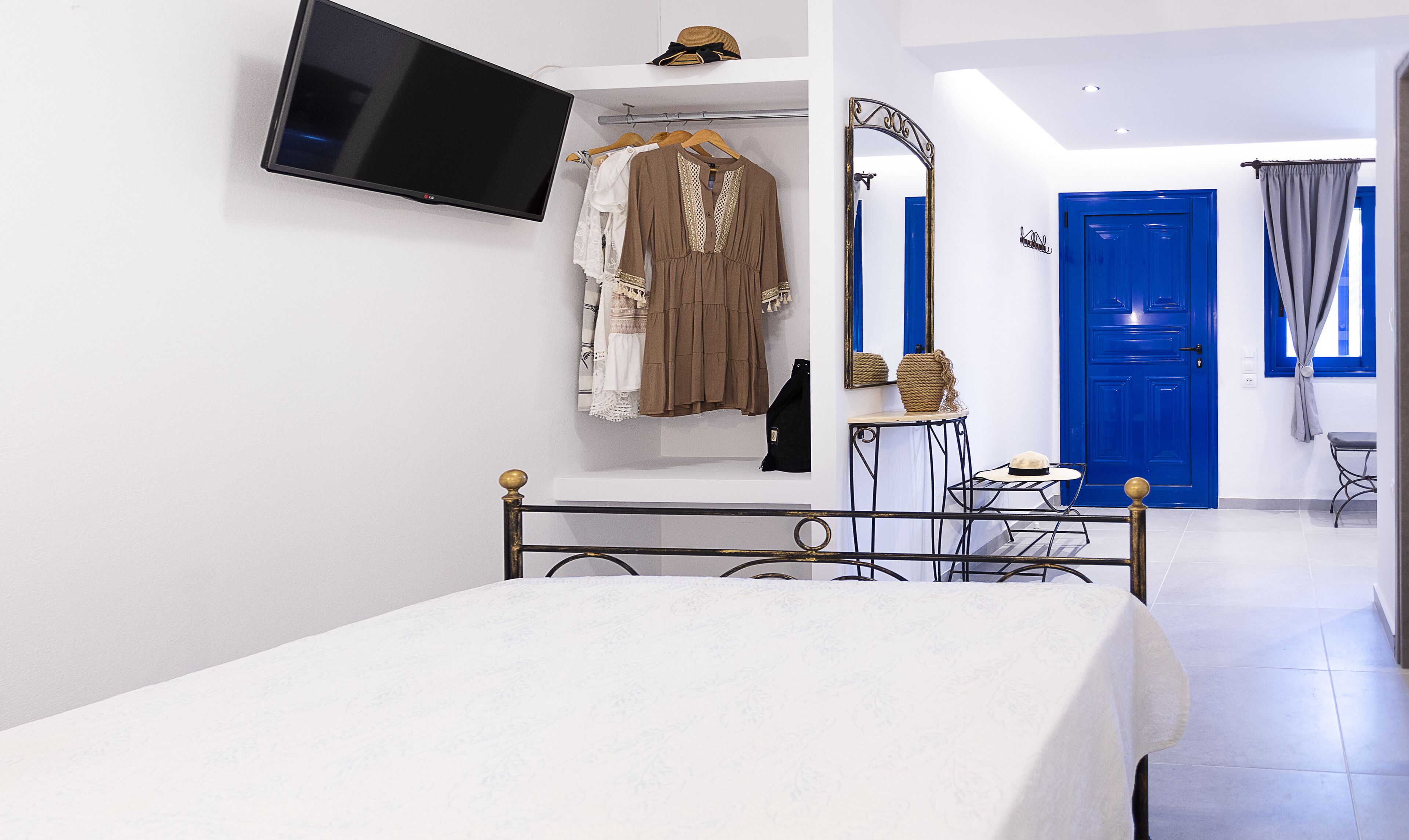 A bedroom with a bed, a TV mounted on the wall, a wardrobe with clothes, a blue door, and a small table with a hat.