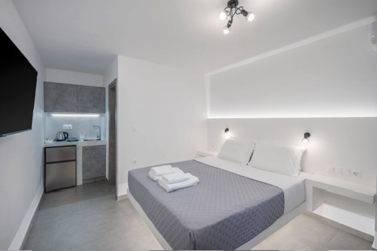 A modern hotel room with a bed, bedside tables, a built-in TV, and a kitchenette.