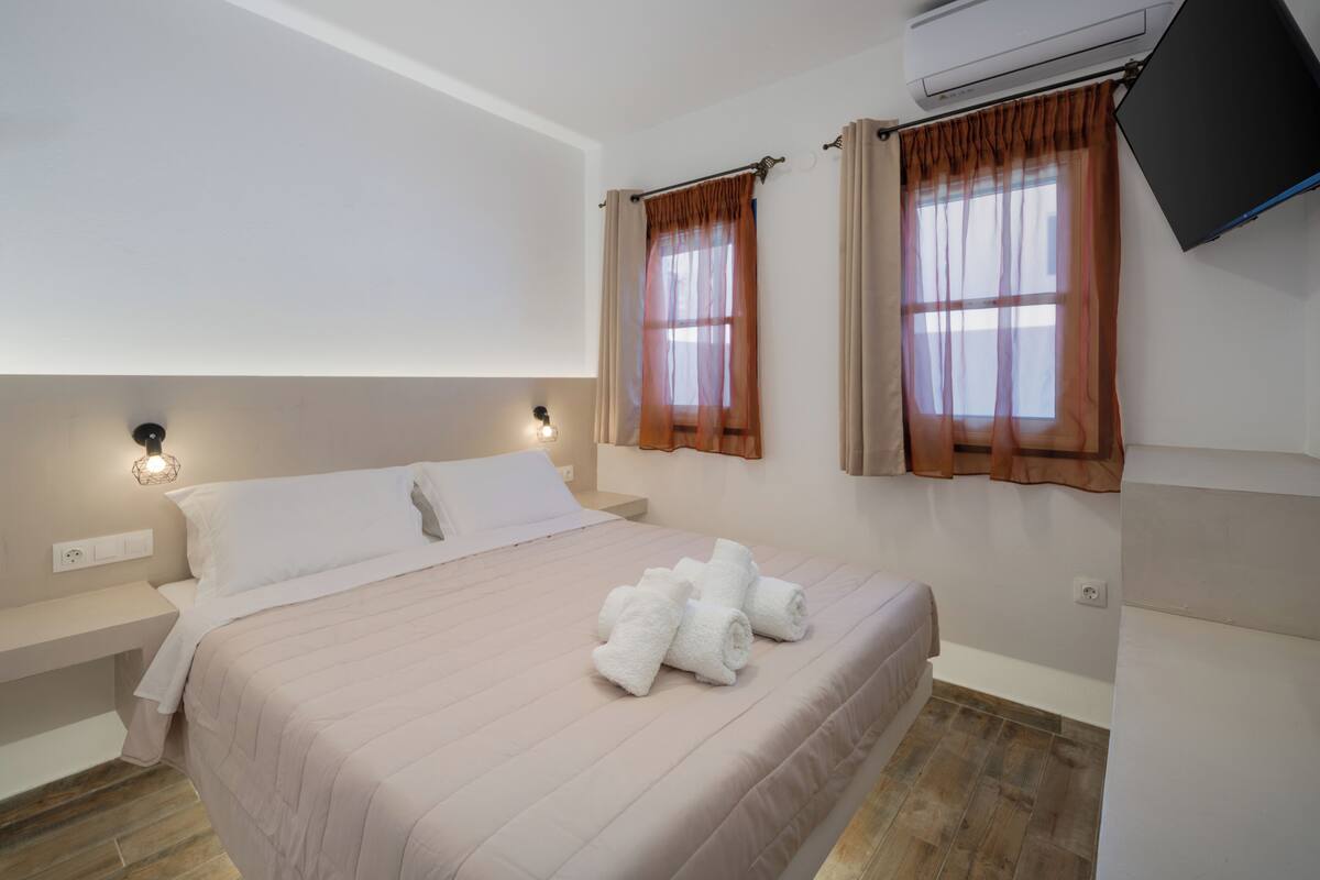 A bedroom with a bed, two windows with curtains, a wall-mounted TV, and a built-in shelf.