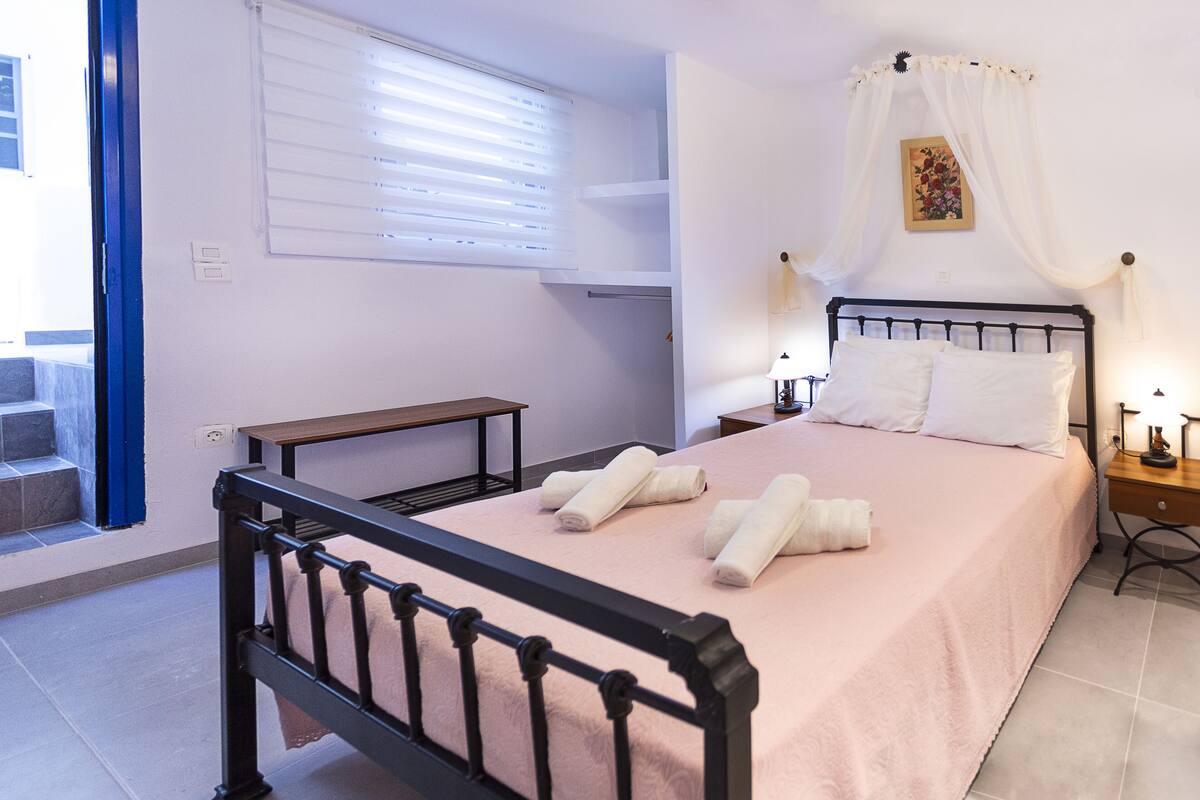 A neatly made bed with pillows and towels, a bedside table with a lamp, a window with blinds, and a small bench.