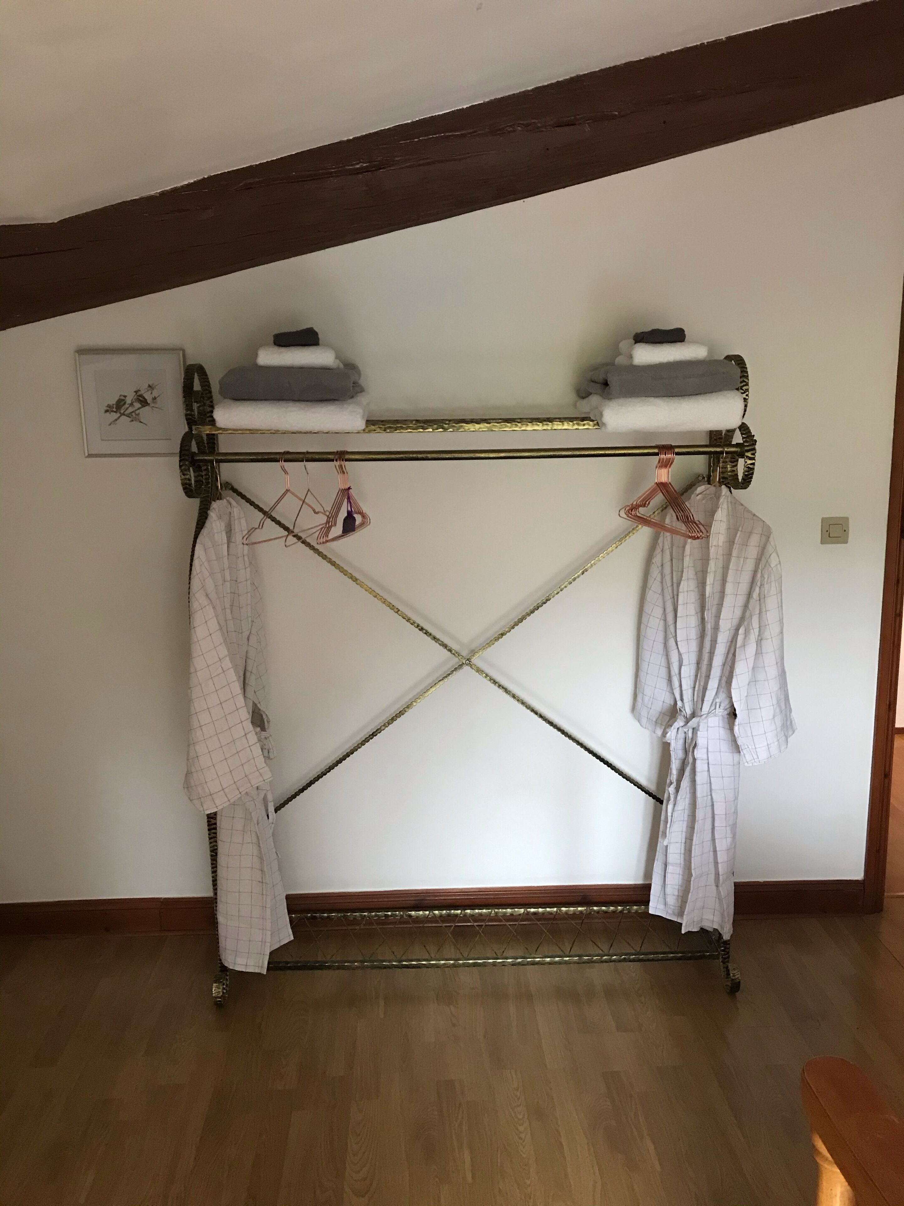 A room with a wooden floor, a white wall, and a ceiling with exposed beams. There is a metal drying rack with two white robes hanging on it.