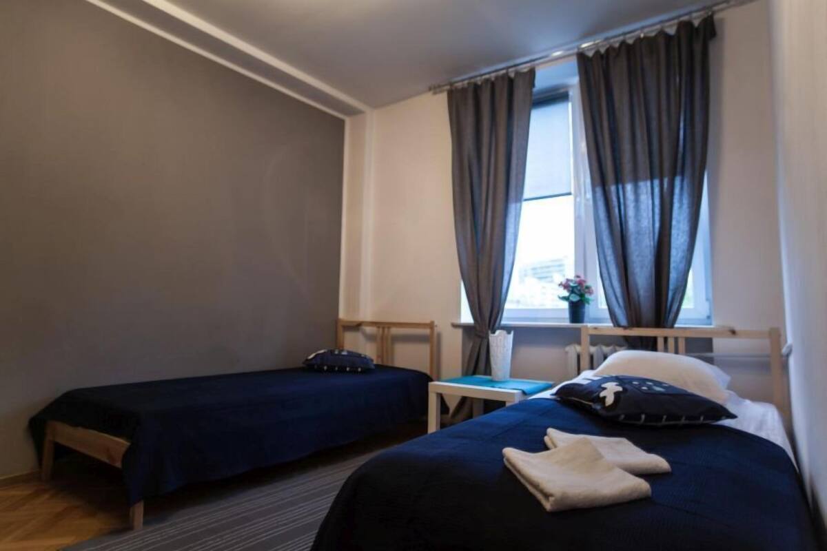 A hotel room with two single beds, a small table, a chair, a window with curtains, and a radiator.