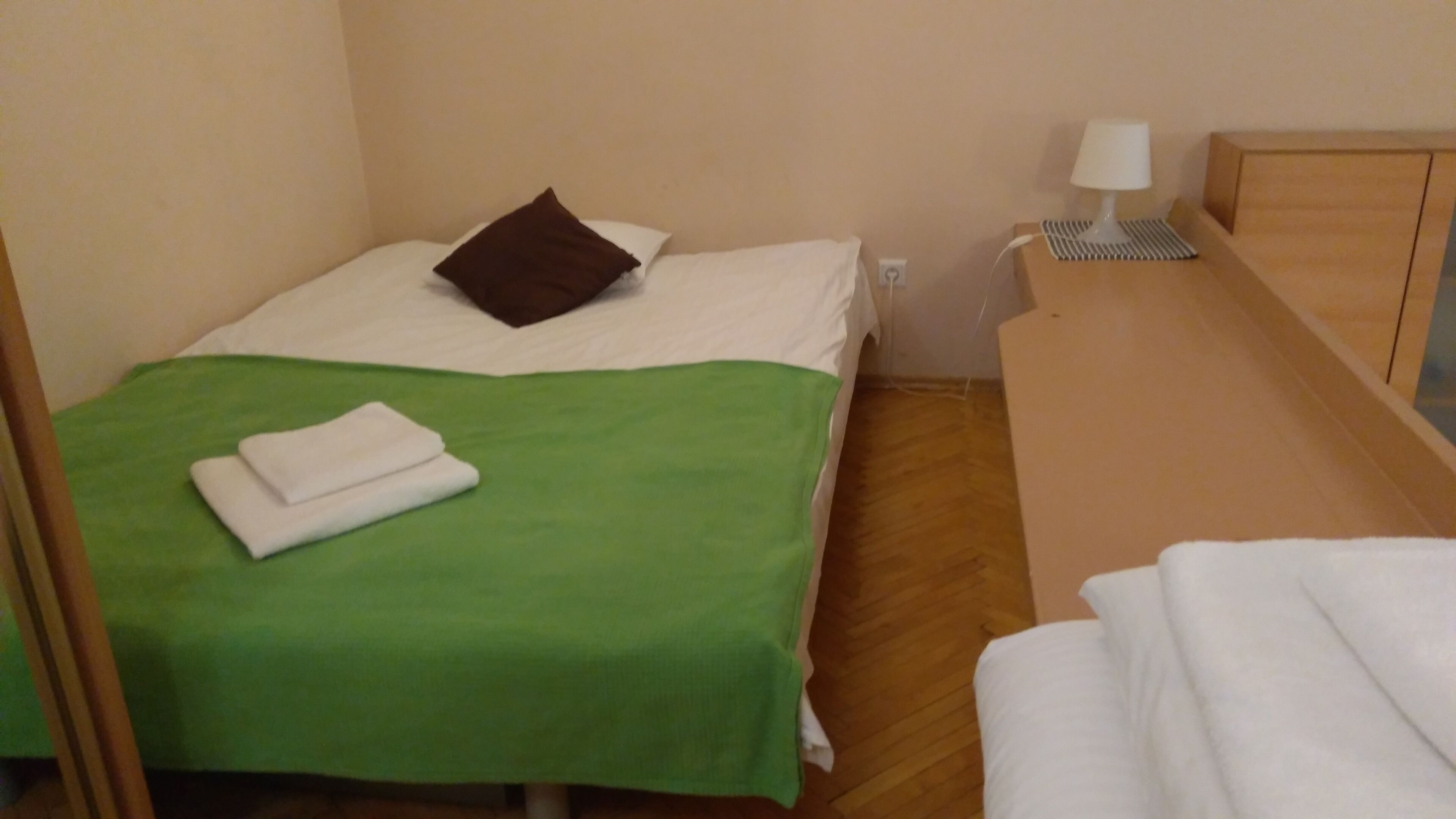 A small, neatly made room with a bed, a bedside table with a lamp, and a wooden nightstand.
