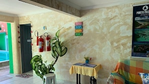 A lobby with a green door, a fire extinguisher, a plant, a small table with a book, and a colorful couch. - Casa Porto Hospedaria (Ipojuca)