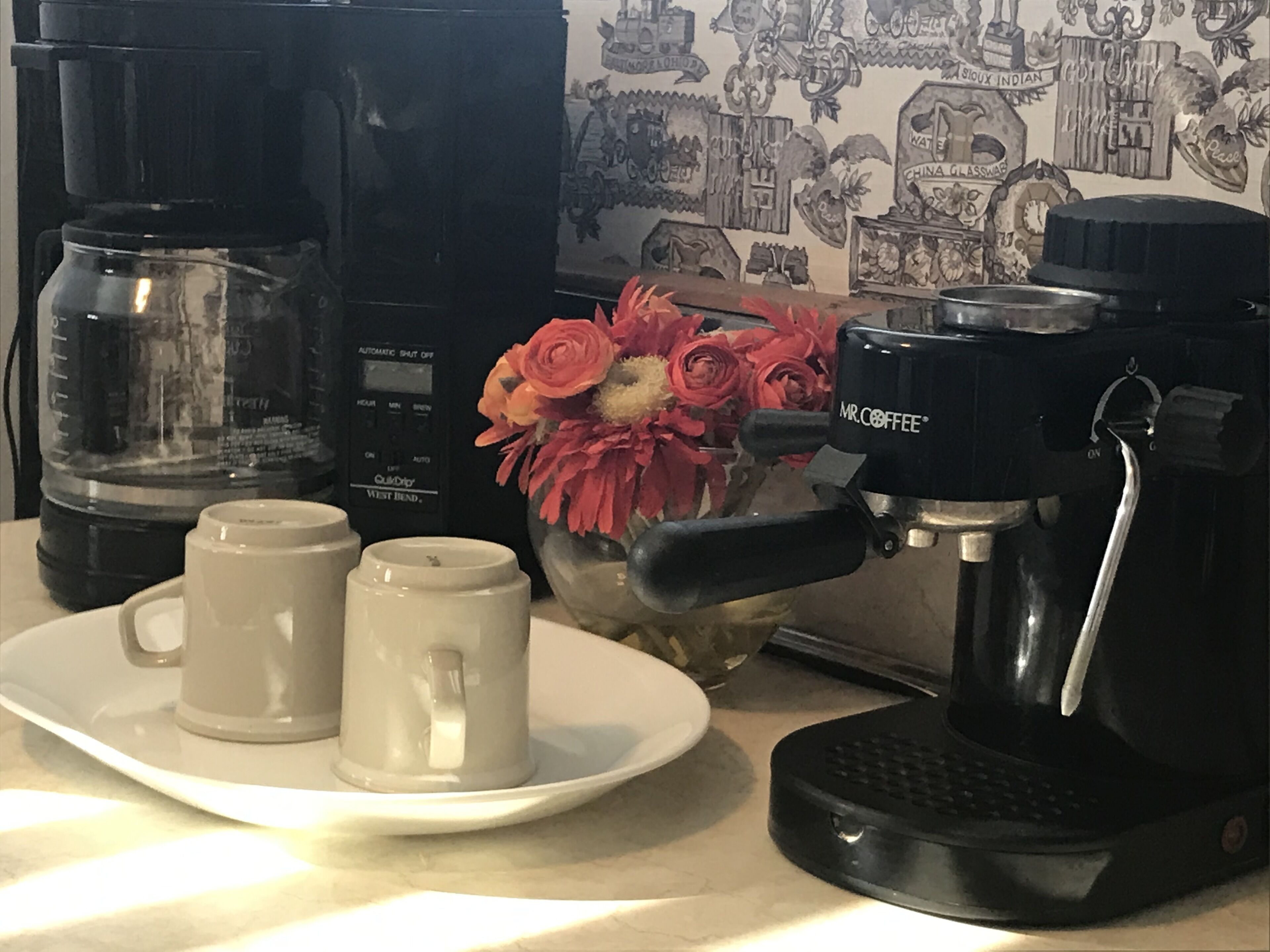 Una cafetera, dos tazas y un florero con flores sobre una mesa.