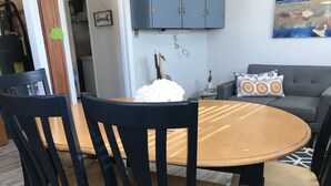 Un comedor con una mesa redonda de madera y sillas negras. Se ve un sofá gris con cojines estampados al fondo.