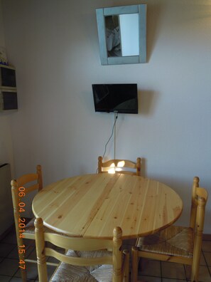 Une table de salle à manger ronde en bois, accompagnée de quatre chaises, un téléviseur à écran plat fixé au mur et une petite étagère sur le côté gauche.