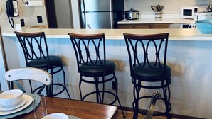 A kitchen with wooden cabinets, a stainless steel refrigerator, a microwave, and a stove. There are three bar stools and a dining table with plates and bowls.