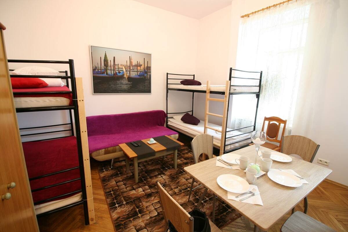 A dormitory room with bunk beds, a sofa, a dining table with plates and cutlery, and a painting of a canal scene.