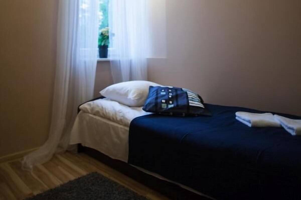 A small, neatly arranged bedroom with a single bed, a window with sheer curtains, a dark-colored rug, and a small potted plant on the windowsill.