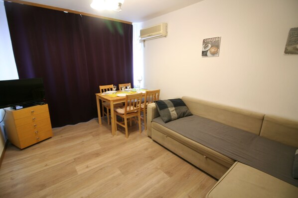 A small, furnished room with a wooden floor, a sofa, a dining table with chairs, a television, and an air conditioning unit.