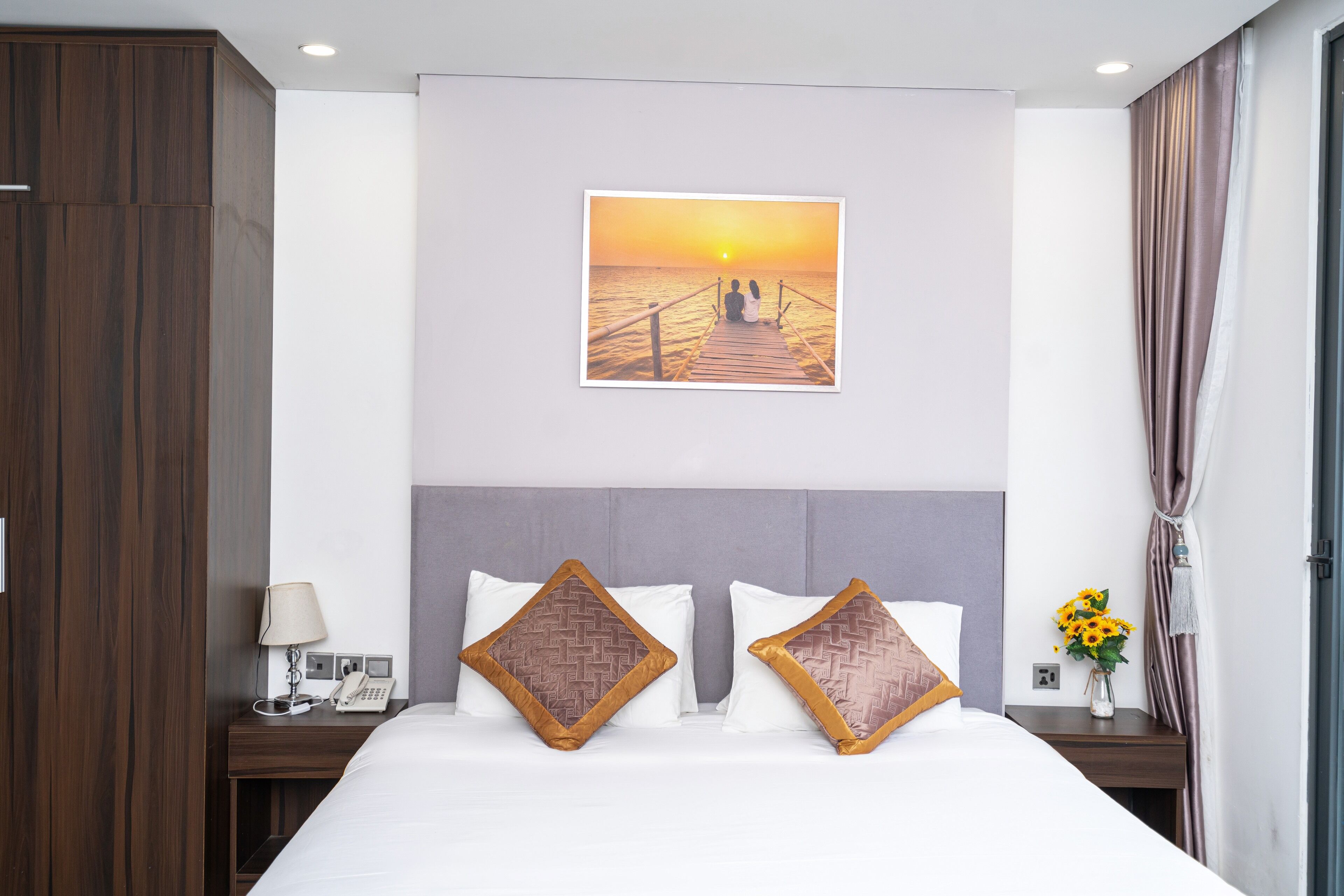 Habitación de hotel con una cama grande, una mesita de noche con lámpara y teléfono, una imagen de un atardecer sobre un muelle, y un florero con flores.