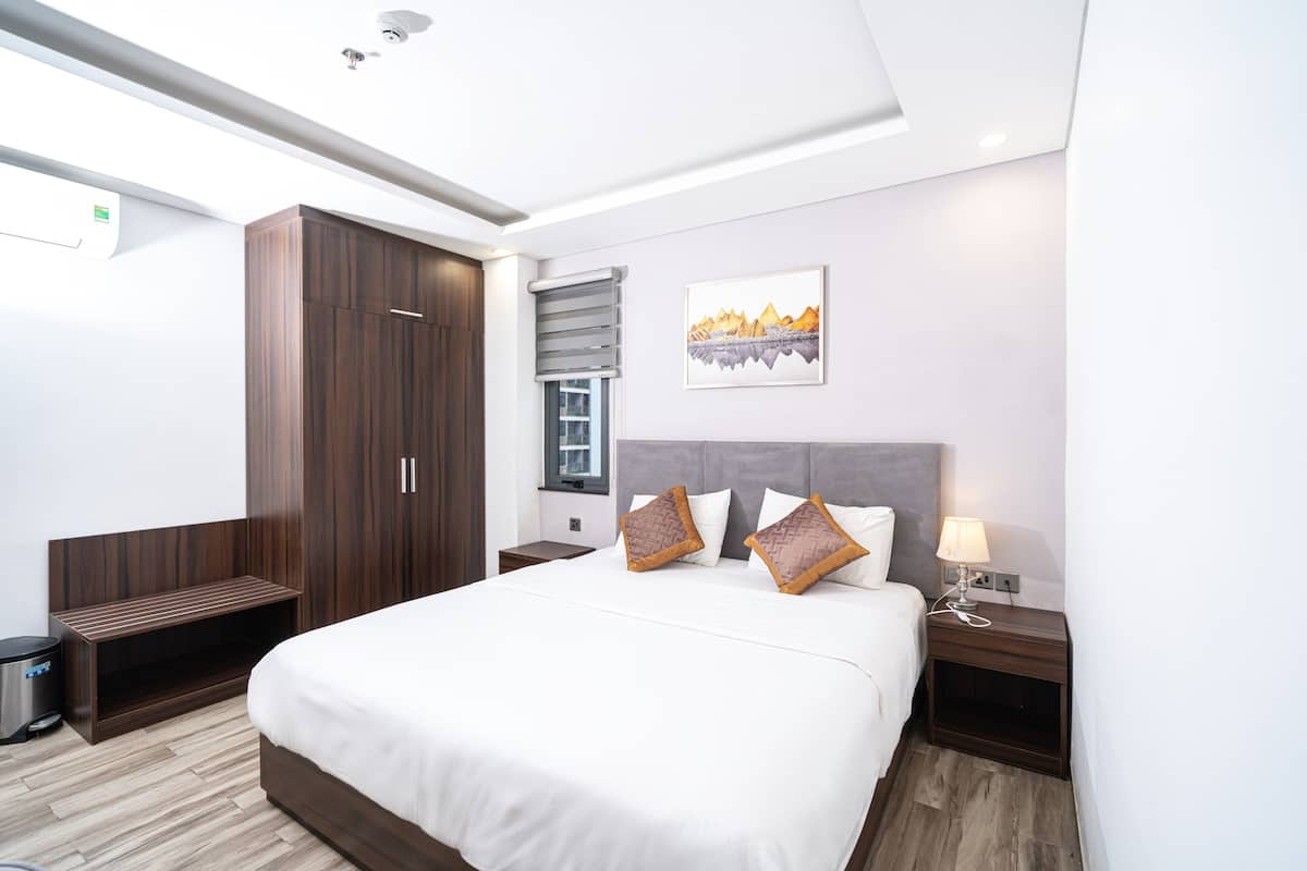 A modern hotel room with a large bed, a wardrobe, a bedside table with a lamp, and a window with blinds.