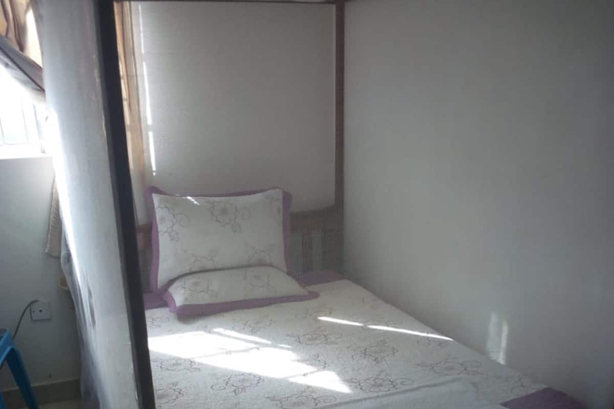 A small, single-bed room with a plain white wall and a window allowing natural light.