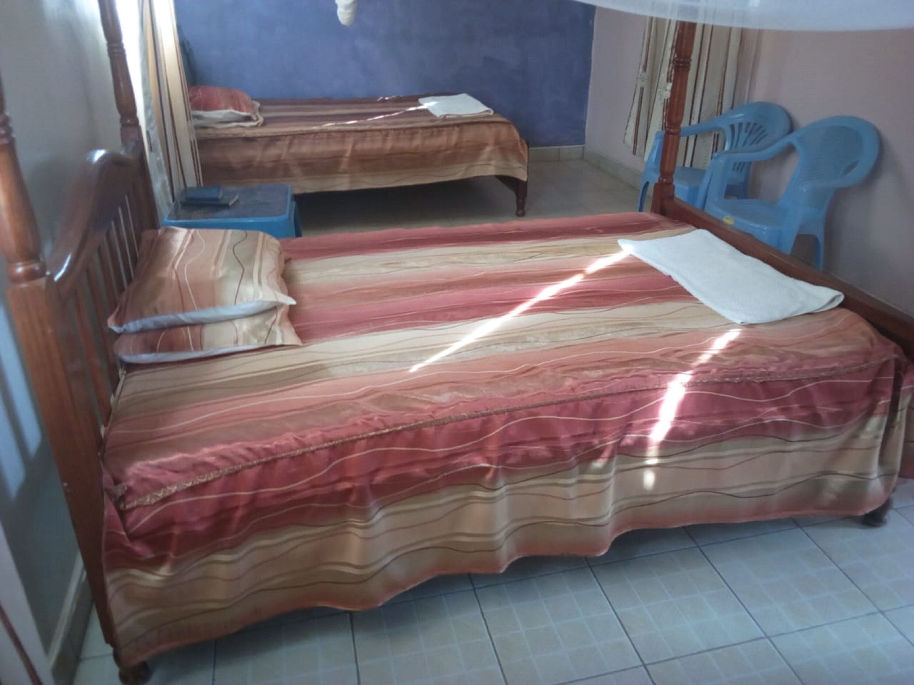 A room with two beds, a wooden chair, a blue plastic chair, and a small table.