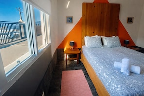 A hotel room with a large bed, a wooden headboard, a nightstand, a window with a view of the street, and a red rug. - Sea Roots "Sea zone" - Amazing sunsets and ocean views (Mosteiros)