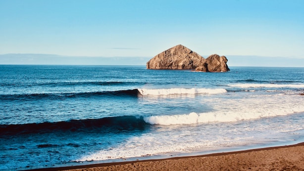 Une plage avec des vagues et une grande formation rocheuse visible au loin.