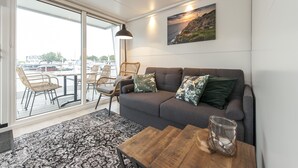A modern living room with a grey sofa, wicker chairs, a wooden coffee table, and a large window with a view of parked cars and greenery outside. - Cozy floating Boatlodge "Mallorca", Max. 4 pers, 2 bedrooms. (Maastricht)