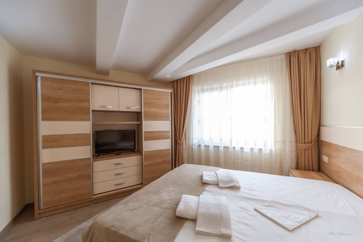 A hotel room with a bed, a TV, a wardrobe, and a large window with curtains.
