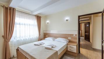 A neatly made bed with a wooden headboard, a bedside table, and a nightstand. There is a window with sheer curtains and a view of a corridor.