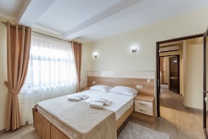 A neatly made bed with a wooden headboard, a bedside table, and a nightstand. There is a window with sheer curtains and a view of a corridor. - Dany Luxury Apartment (Pitesti)