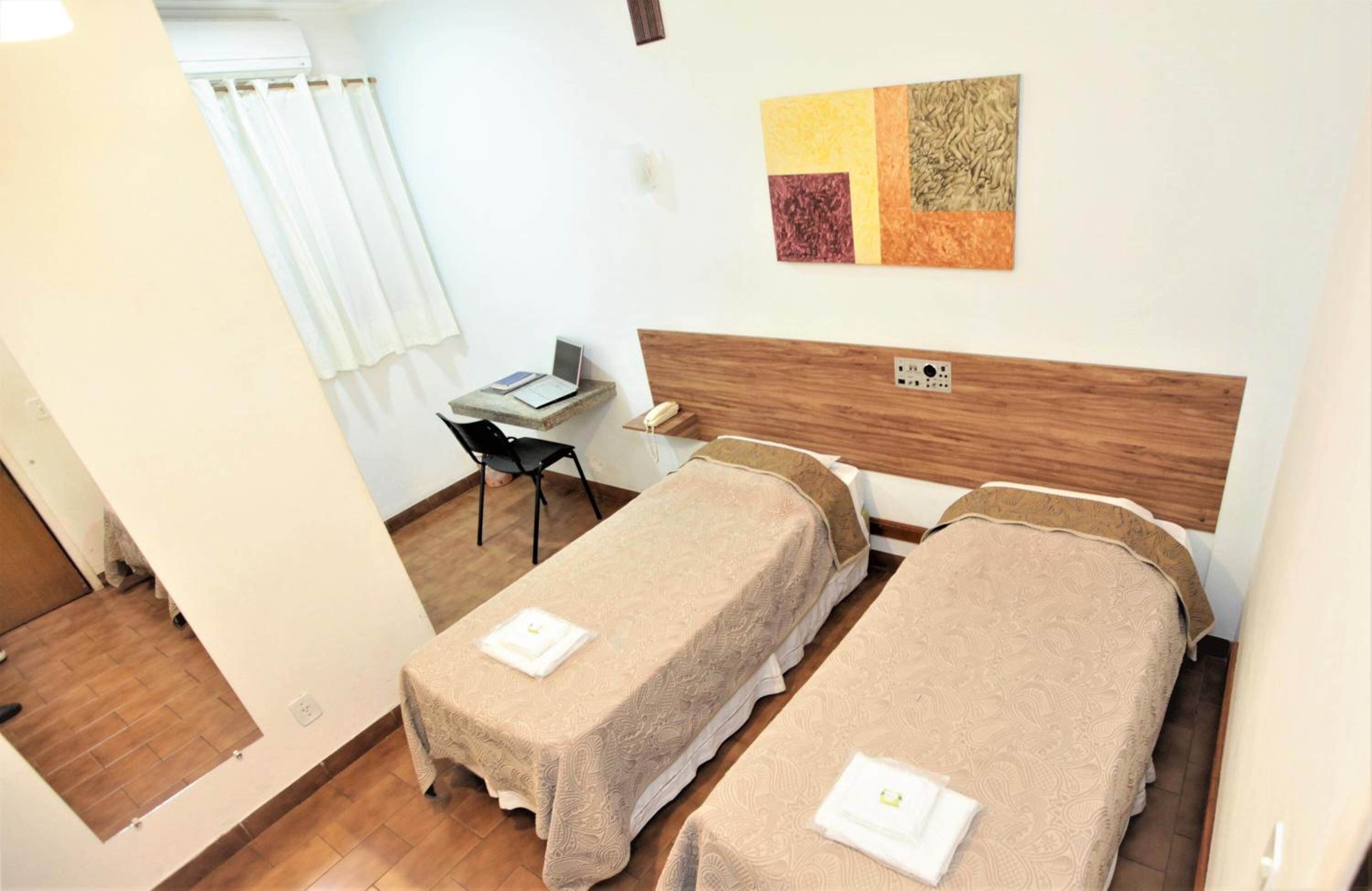 A hotel room with two beds, a desk with a laptop, a chair, and a wall art piece.