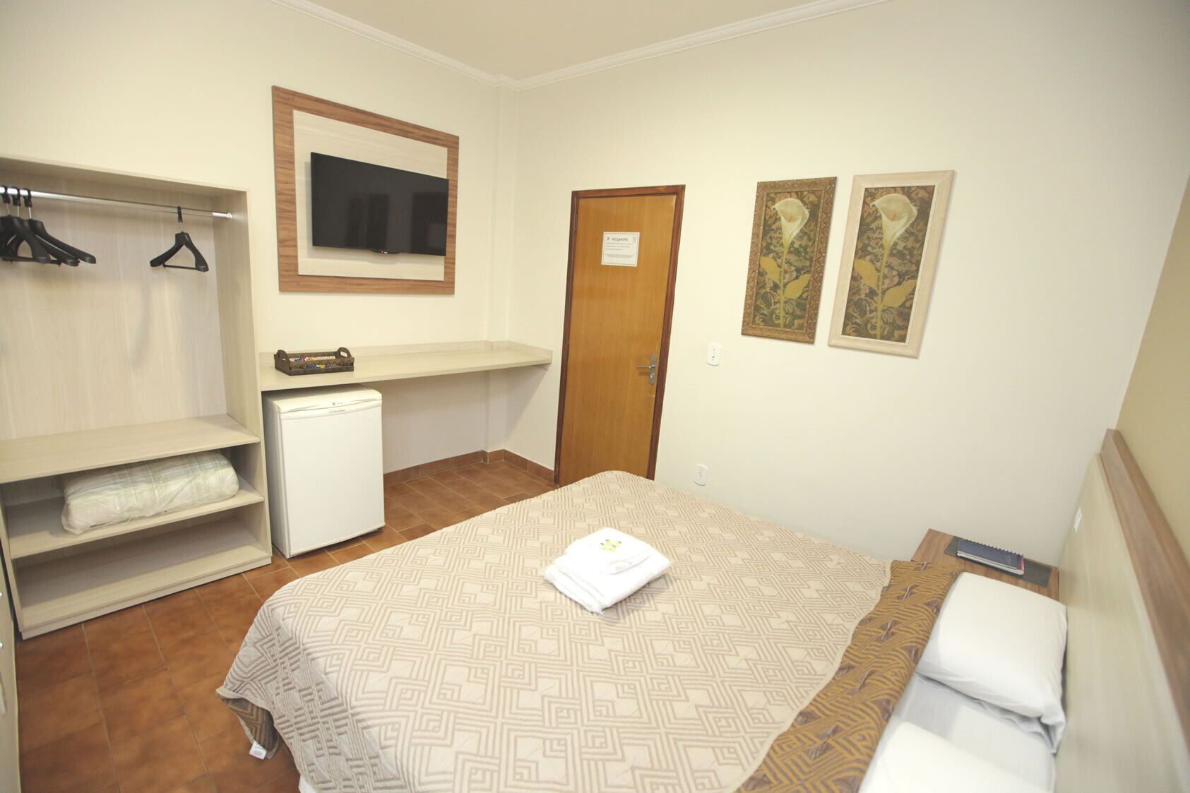 Quarto compacto com cama, escrivaninha pequena, guarda-roupa e televisão.