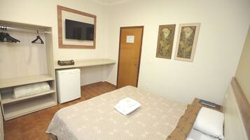 Quarto compacto com cama, escrivaninha pequena, guarda-roupa e televisão.