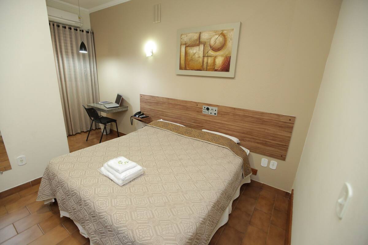 Una habitación de hotel con cama, escritorio, silla, un cuadro en la pared y cortina.