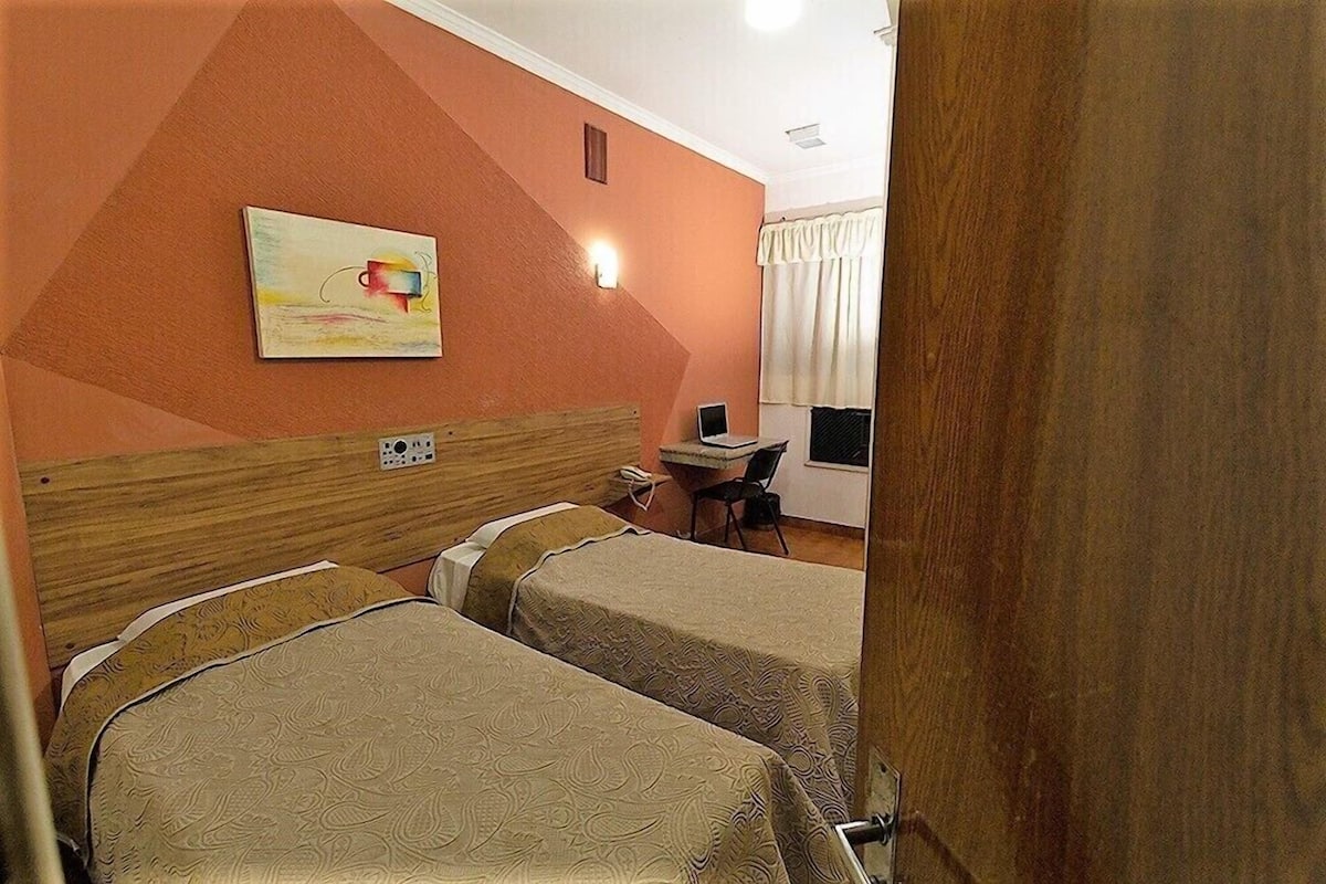 Habitación de hotel con dos camas, un escritorio, una silla, un cuadro en la pared y una ventana con cortinas.