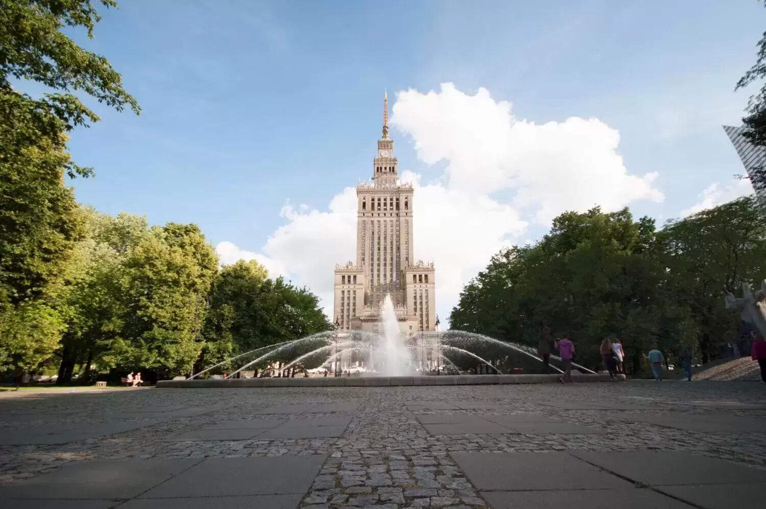 Um edifício grande com uma torre imponente, rodeado por uma fonte e árvores.