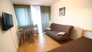 A room with a wooden herringbone floor, a brown sofa with a patterned pillow, a small desk with a chair, a television mounted on the wall, and a poster on the wall. - Apartamenty Varsovie Sliska (Warsaw)