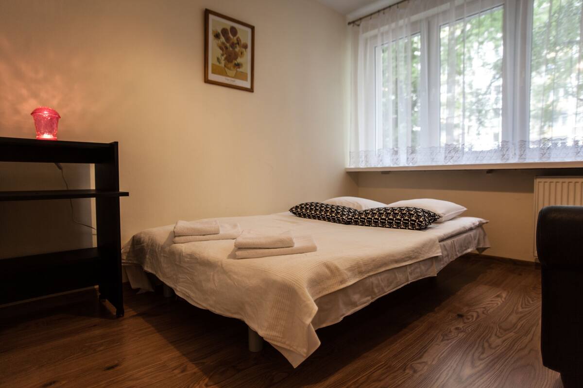 Quarto com uma cama, uma mesa de cabeceira com um objeto vermelho, uma janela com cortinas brancas e um quadro na parede.