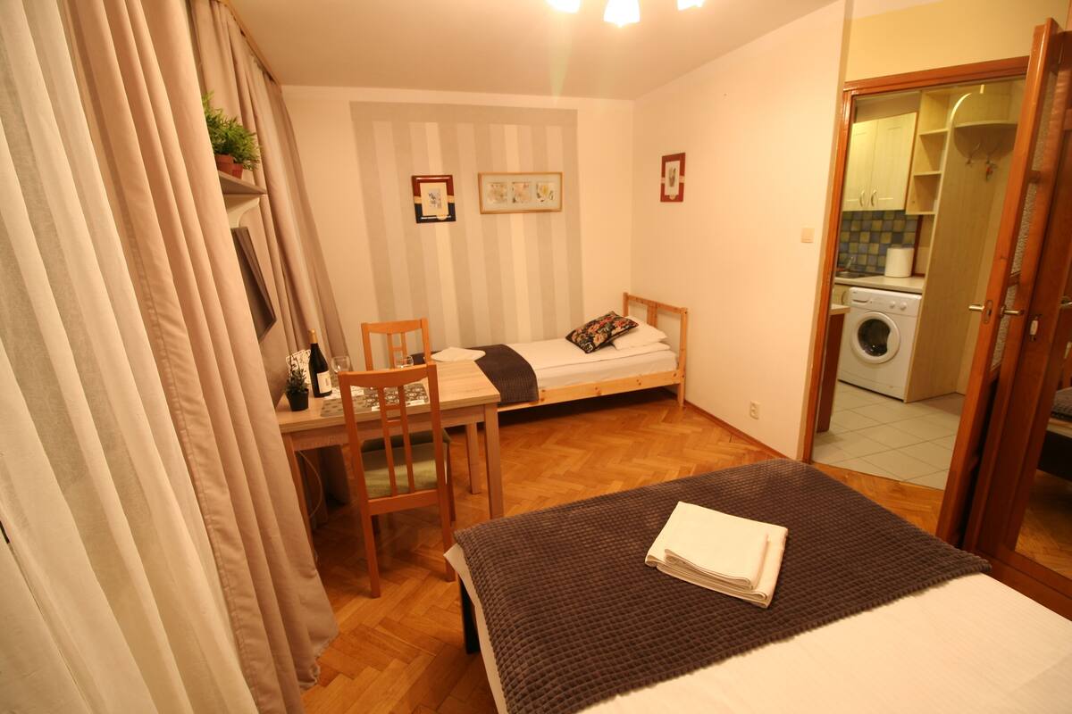 Quarto compacto com uma cama de solteiro, uma pequena escrivaninha, uma cadeira e uma máquina de lavar no quarto ao lado.
