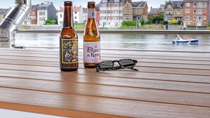 Twee bierflesjes en een zonnebril op een houten tafel, met uitzicht op een gracht en huizen op de achtergrond.