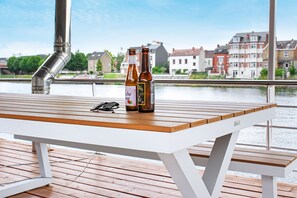 A wooden table with two bottles of beer and a pair of sunglasses on a dock by a river, with buildings in the background.