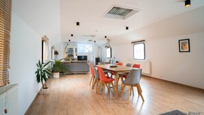 Een ruime woonkamer met een houten vloer, een eettafel met stoelen en een bank. Er staan potplanten en er is een radiator aan de muur.